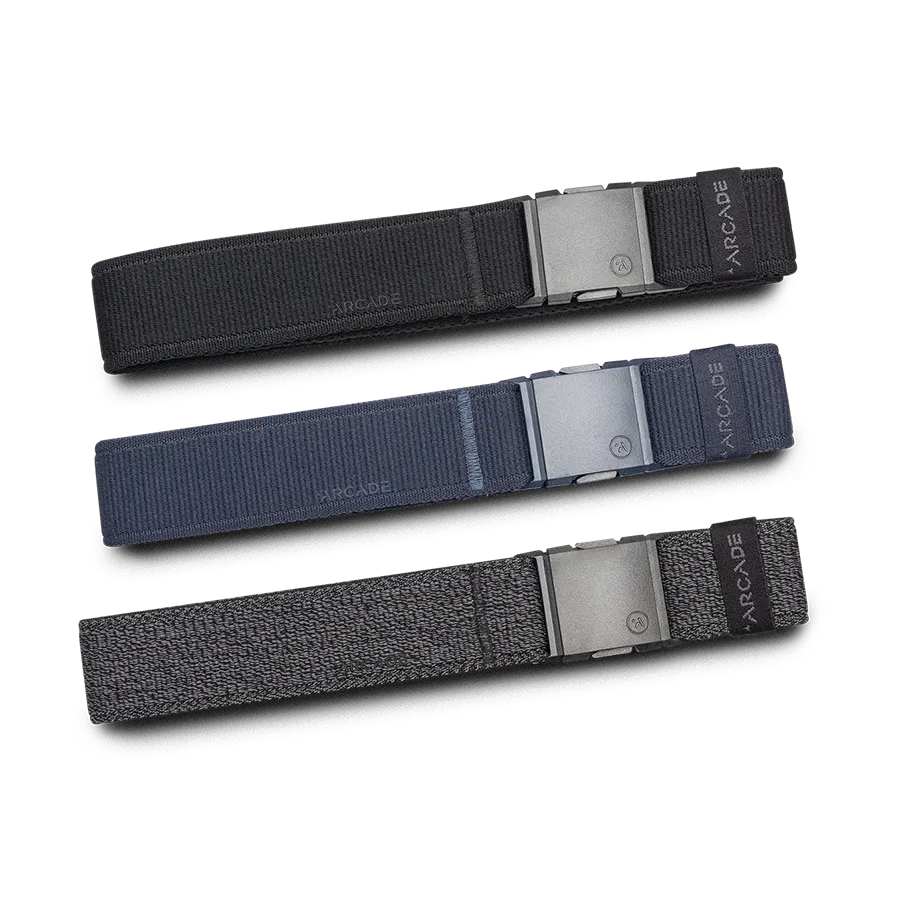 Three fabric belts in black, navy blue, and dark gray are laid out parallel to each other. Each belt features a matte gray buckle and a small label with the brand name ARCADE.