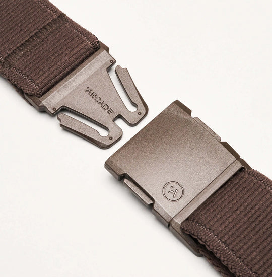Close-up of the brown Atlas belt by Arcade Belts, crafted from stretch webbing. The open metallic buckle displays two pieces—one engraved with ARCADE and the other featuring a small smiling face logo.