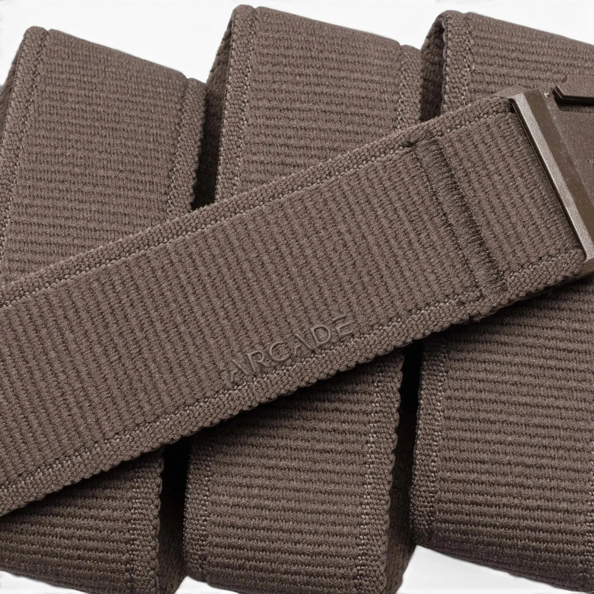 A close-up of the Atlas belt by Arcade Belts, featuring brown woven stretch webbing, a low-profile metal buckle, and a subtly embossed ARCADE logo on the folded strap—highlighting the brand’s signature style.