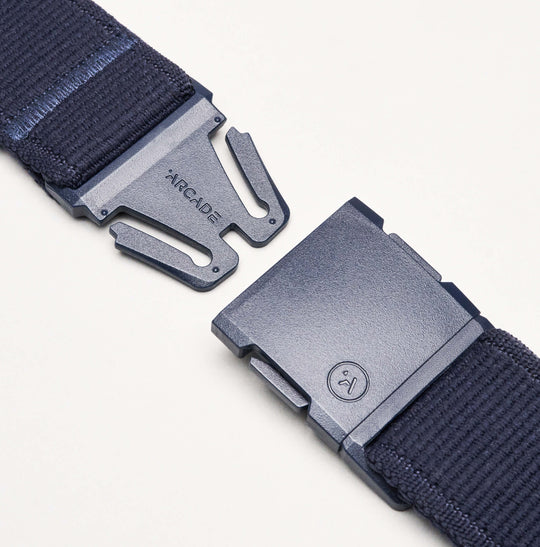 Close-up of the Atlas Elastic Stretch Belt by Arcade Belts in dark blue, featuring a metal buckle engraved with ARCADE. The partially unclasped buckle reveals the interlocking mechanism, emphasizing the belts stylish and functional travel-ready design.