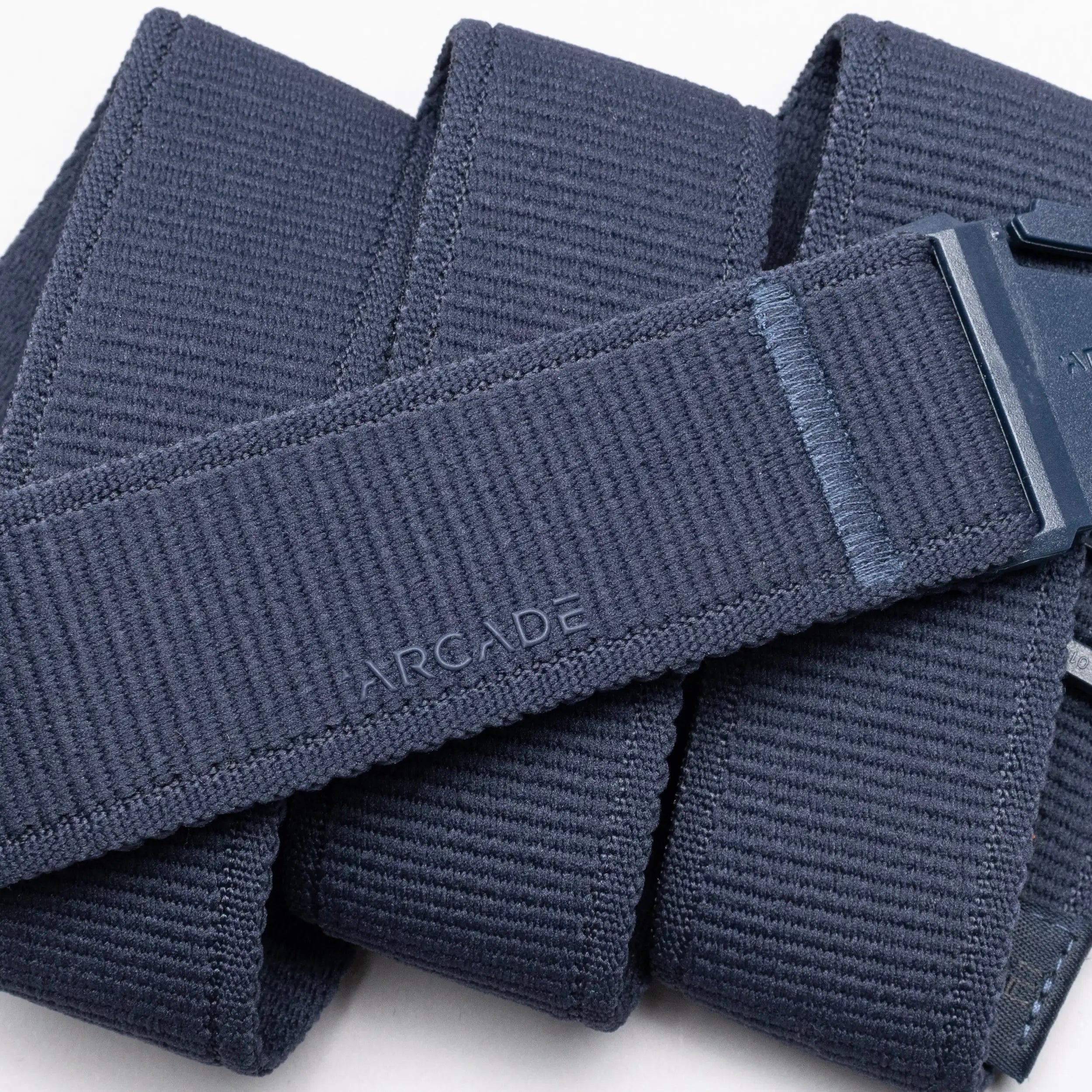 A close-up image features the Atlas belt by Arcade Belts, a navy blue elastic stretch belt with a plastic buckle and subtle ARCADE branding. Neatly folded, it’s perfect for travelers seeking comfort and a low-profile design.