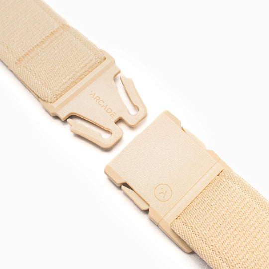 A close-up of the beige Atlas Elastic Stretch Belt by Arcade Belts, showing its plastic buckle slightly open to display the fastening mechanism. ARCADE is engraved on one side. The background is white.