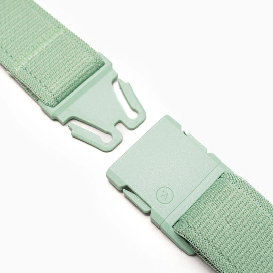 A close-up of the Arcade Belts Atlas shows its green fabric strap with an unfastened plastic buckle, featuring a minimalist smiley face icon, lying on a white background.