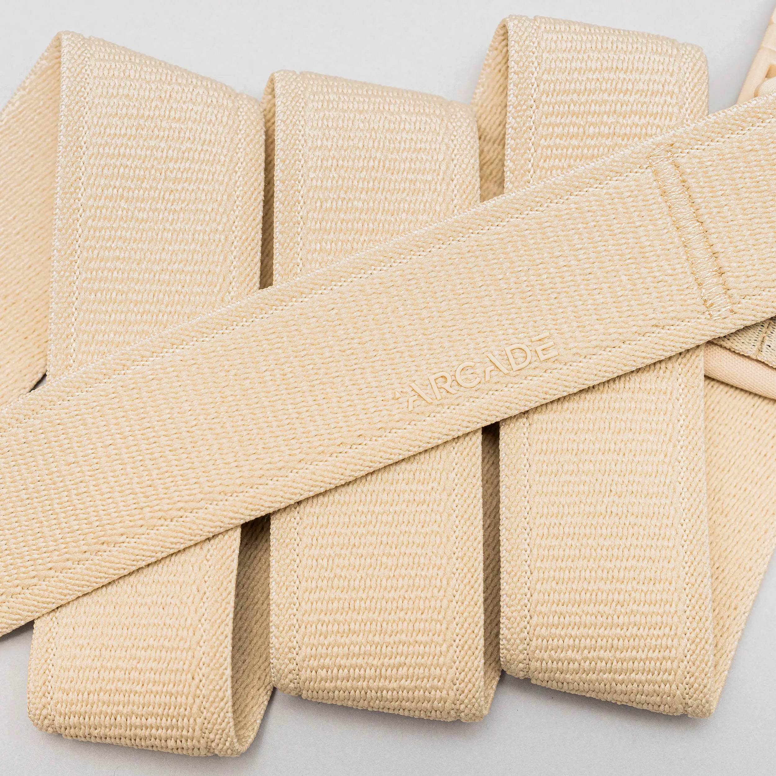 A close-up of beige woven fabric straps in a crisscross pattern features ARCADE embossed on the center, highlighting the Atlas elastic stretch belt by Arcade Belts—an ideal choice for travel.