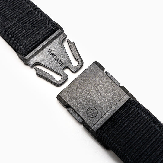 Close-up of the Atlas belt by Arcade Belts, a black elastic stretch belt with a metal quick-release buckle—partially unclasped, ARCADE engraved on one side. White background.
