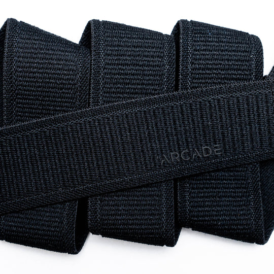 Close-up of several black Atlas straps by Arcade Belts stacked together, with one crossing diagonally and subtly embossed with ARCADE, showcasing the quality of this elastic stretch belt.