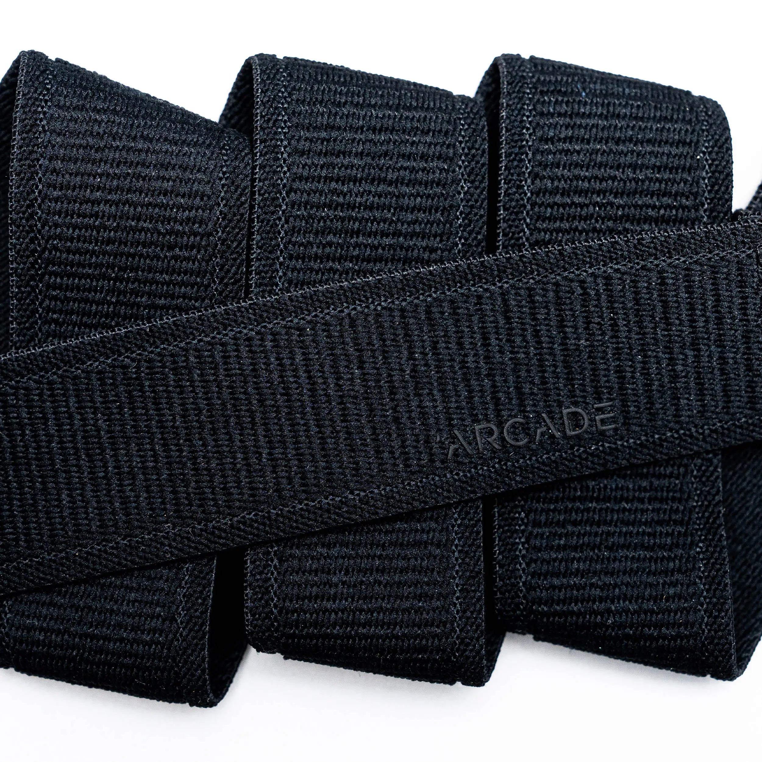 Close-up of several black Atlas straps by Arcade Belts stacked together, with one crossing diagonally and subtly embossed with ARCADE, showcasing the quality of this elastic stretch belt.