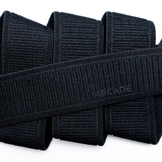 Close-up of several black Atlas straps by Arcade Belts stacked together, with one crossing diagonally and subtly embossed with ARCADE, showcasing the quality of this elastic stretch belt.