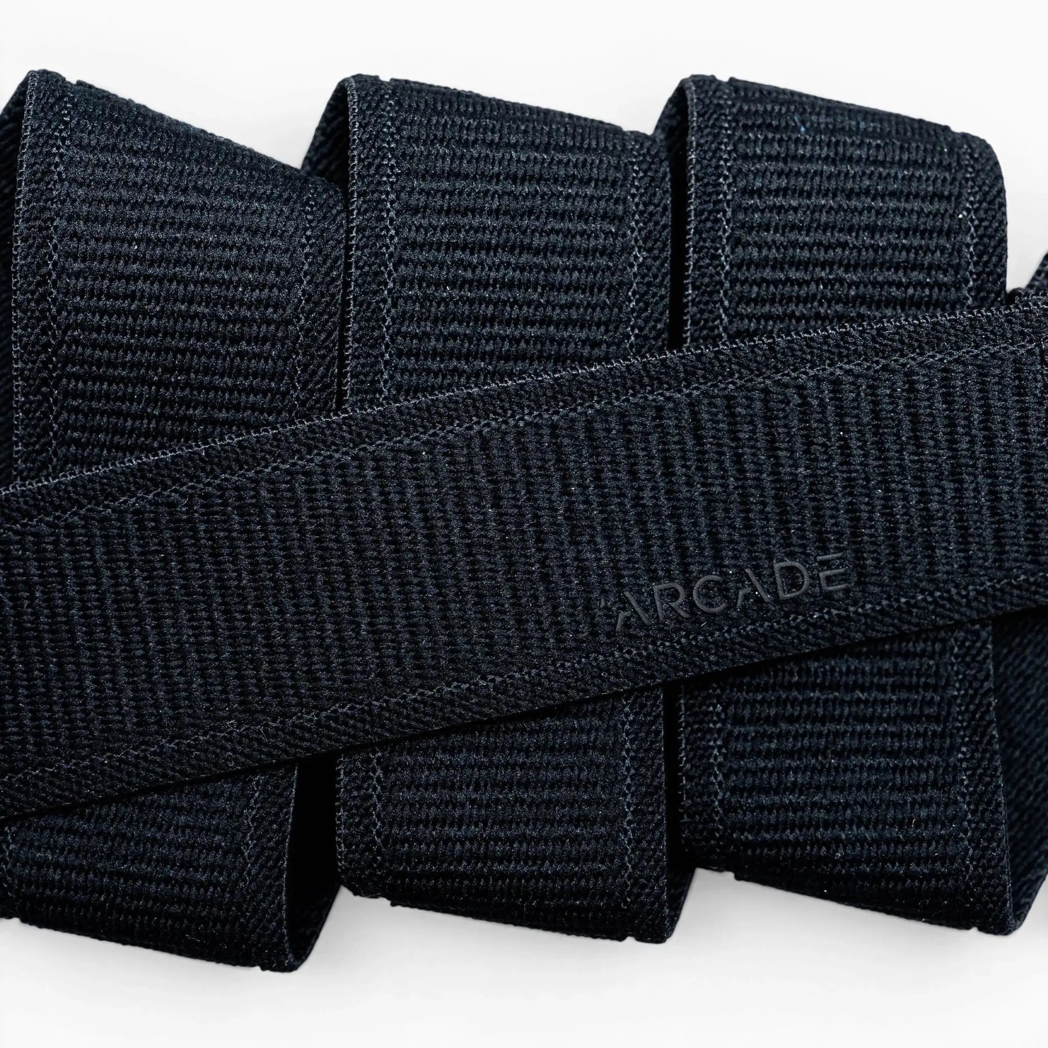 Close-up of several black Atlas straps by Arcade Belts stacked together, with one crossing diagonally and subtly embossed with ARCADE, showcasing the quality of this elastic stretch belt.