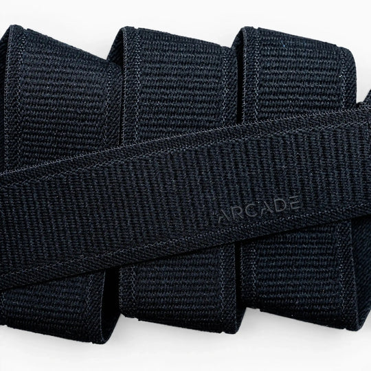 Close-up of several black Atlas straps by Arcade Belts stacked together, with one crossing diagonally and subtly embossed with ARCADE, showcasing the quality of this elastic stretch belt.