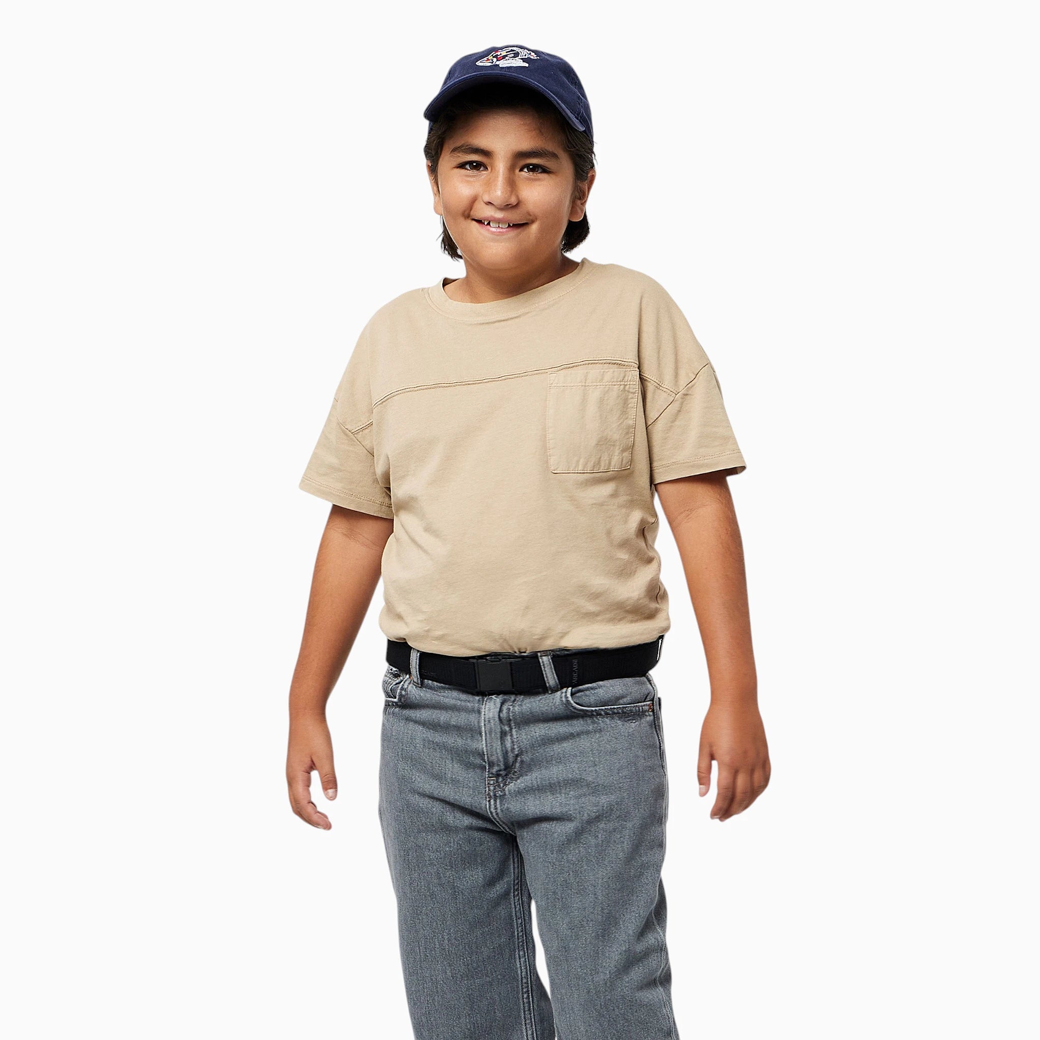 A smiling child wearing a navy baseball cap, beige t-shirt, black belt, and gray jeans stands against a plain white background, proudly showing off their Arcade Belts A2 Youth Kit—a comfortable kids harness that’s machine-washable for easy care.