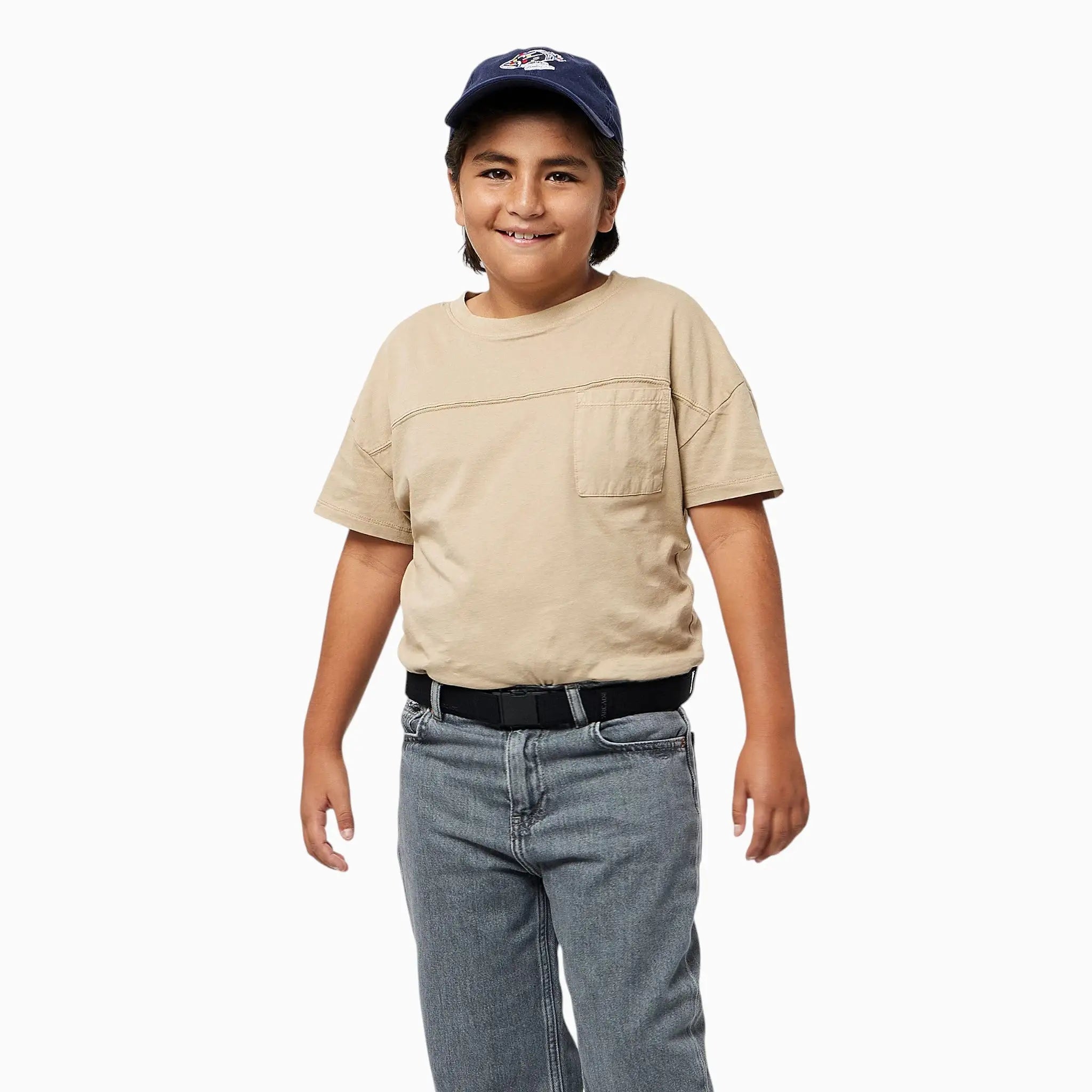 A smiling child wearing a navy baseball cap, beige t-shirt, black belt, and gray jeans stands against a plain white background, proudly showing off their Arcade Belts A2 Youth Kit—a comfortable kids harness that’s machine-washable for easy care.