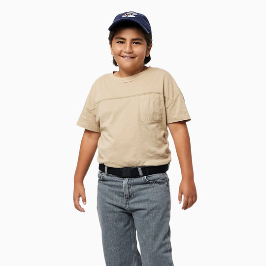 A smiling child wearing a navy baseball cap, beige t-shirt, black belt, and gray jeans stands against a plain white background, proudly showing off their Arcade Belts A2 Youth Kit—a comfortable kids harness that’s machine-washable for easy care.