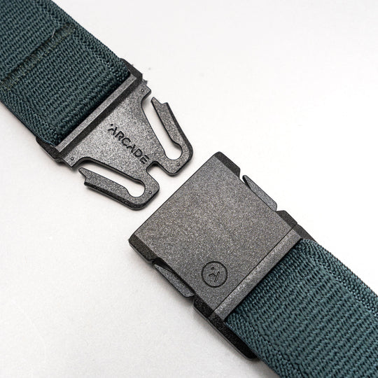 A close-up of the green Atlas travel belt by Arcade Belts, showing a black metal buckle engraved with ARCADE and featuring a two-prong locking mechanism. The belt is partially unfastened.