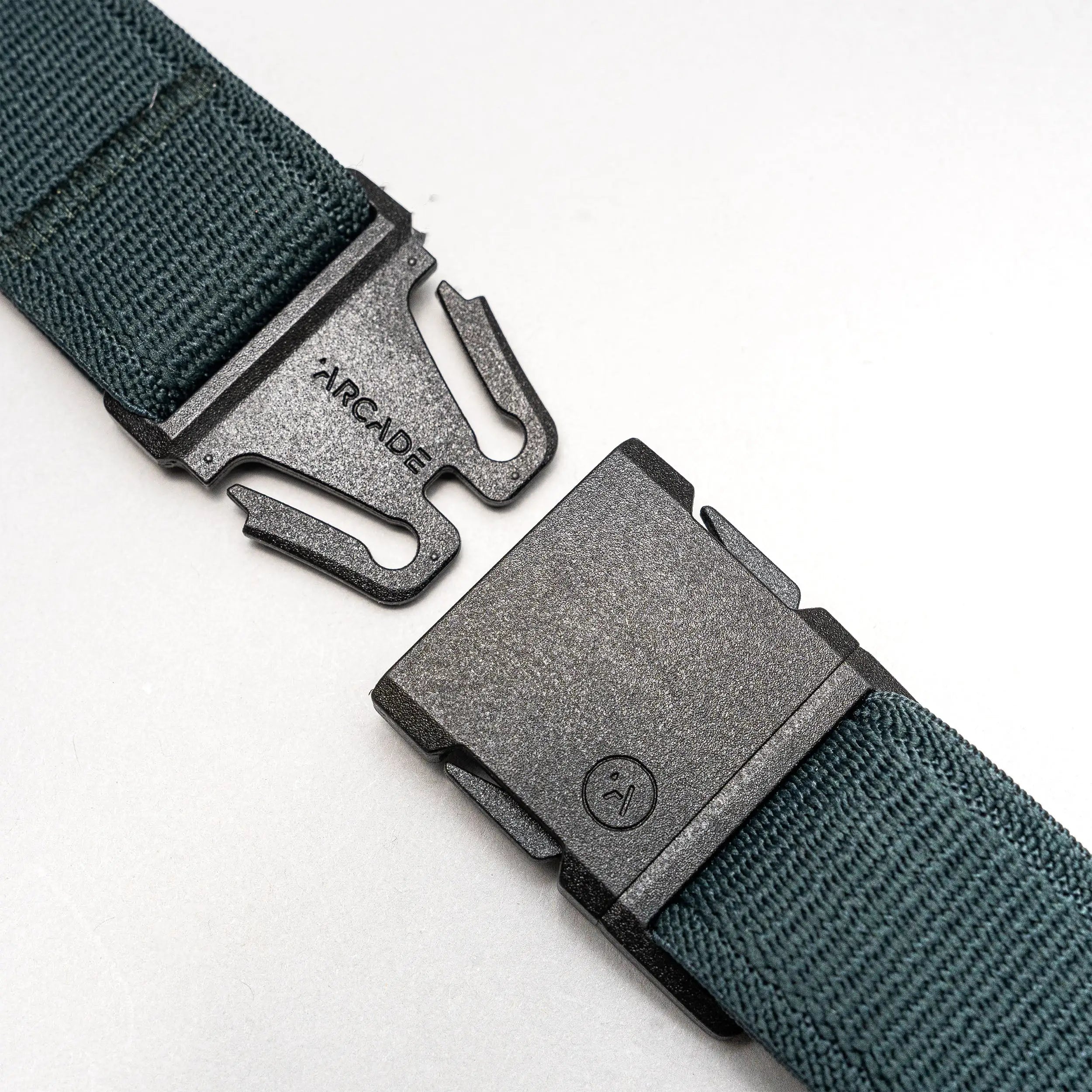 A close-up of the green Atlas travel belt by Arcade Belts, showing a black metal buckle engraved with ARCADE and featuring a two-prong locking mechanism. The belt is partially unfastened.