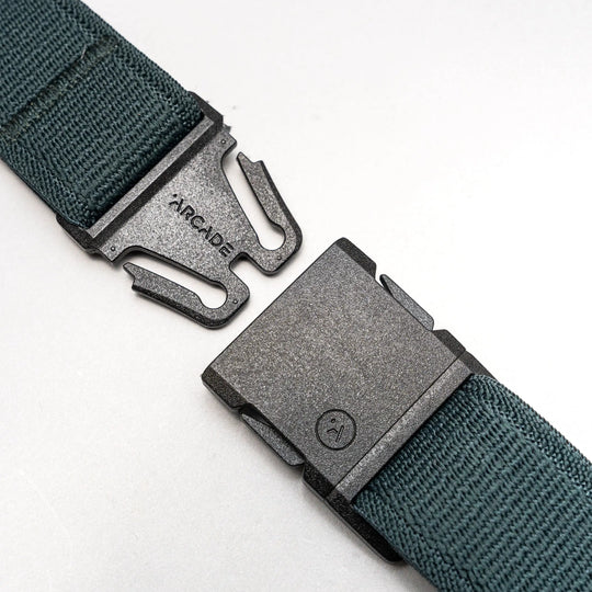 A close-up of the green Atlas travel belt by Arcade Belts, showing a black metal buckle engraved with ARCADE and featuring a two-prong locking mechanism. The belt is partially unfastened.