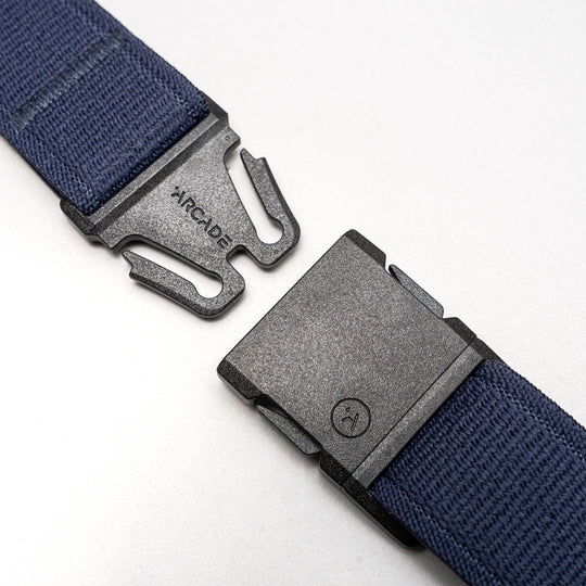 A close-up of a black plastic buckle with Arcade Belts branding on one side, attached to the navy blue Atlas elastic stretch belt, shown unbuckled against a light gray background.