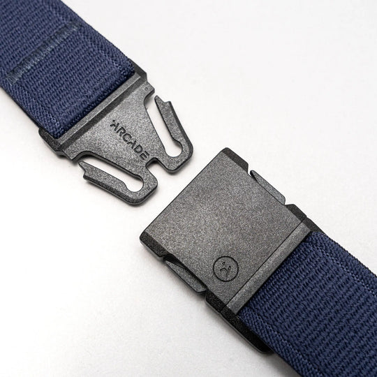 A close-up of a black plastic buckle with Arcade Belts branding on one side, attached to the navy blue Atlas elastic stretch belt, shown unbuckled against a light gray background.
