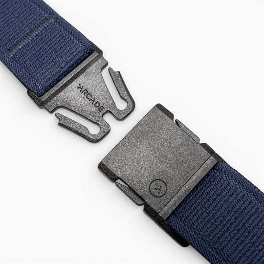A close-up of a black plastic buckle with Arcade Belts branding on one side, attached to the navy blue Atlas elastic stretch belt, shown unbuckled against a light gray background.