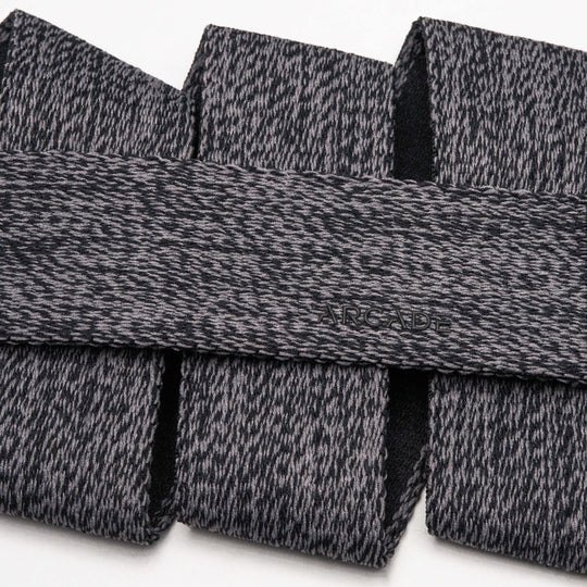 A close-up of the Arcade Belts Atlas, featuring gray and black textured fabric with a subtle embossed logo and a sleek plastic buckle, arranged in overlapping folds on a white background.