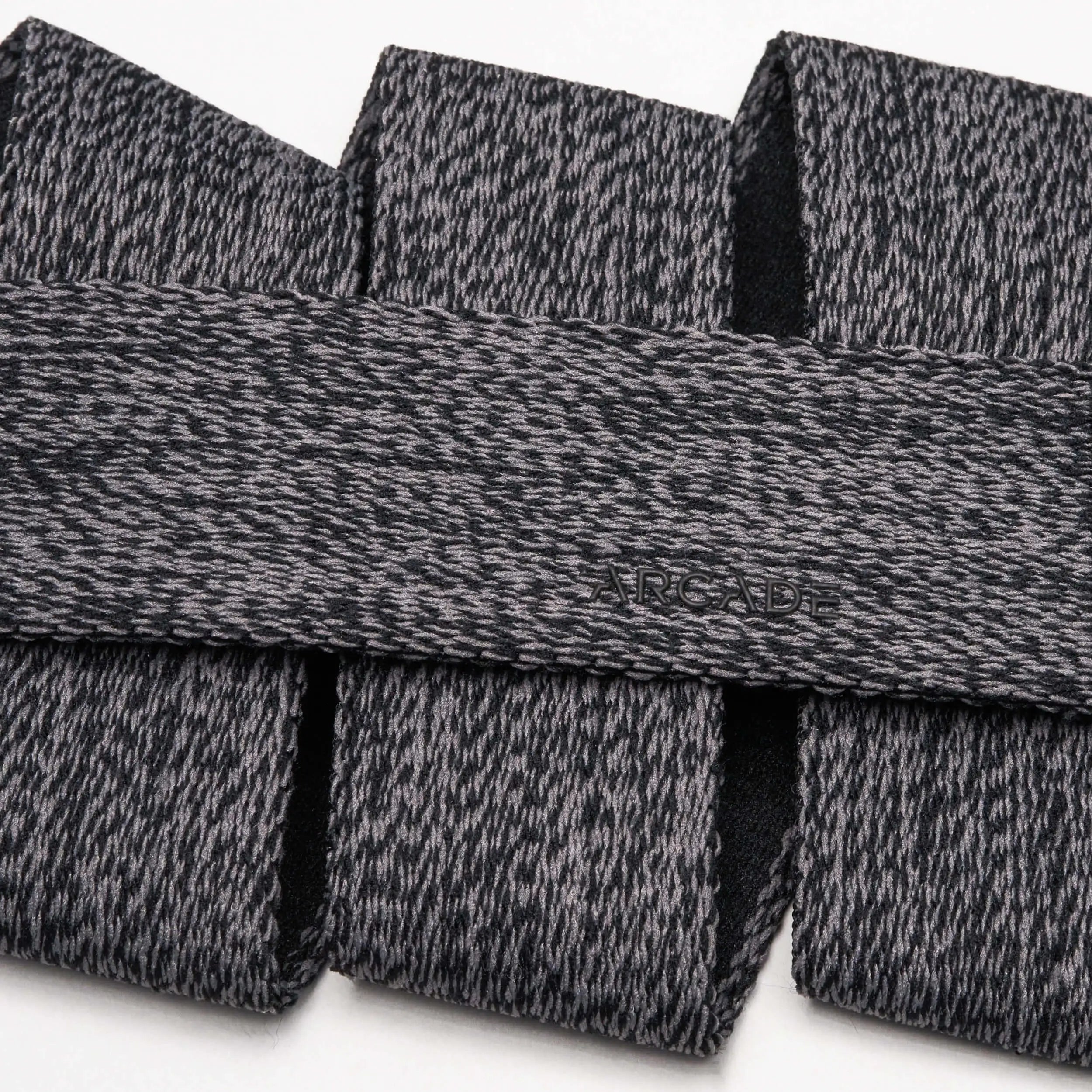 A close-up of the Arcade Belts Atlas, featuring gray and black textured fabric with a subtle embossed logo and a sleek plastic buckle, arranged in overlapping folds on a white background.