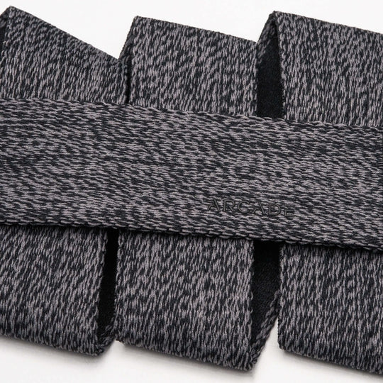 A close-up of the Arcade Belts Atlas, featuring gray and black textured fabric with a subtle embossed logo and a sleek plastic buckle, arranged in overlapping folds on a white background.