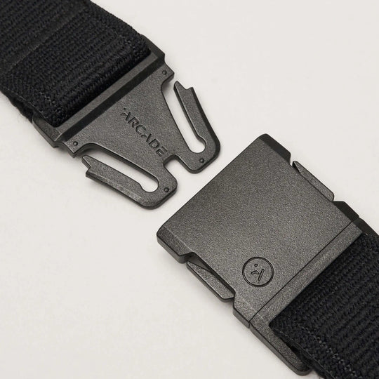 A close-up of the Atlas elastic stretch belt by Arcade Belts in black, featuring a plastic quick-release buckle. The unfastened low-profile buckle shows the Arcade Belts brand name on one side and a small circular face logo on the other.