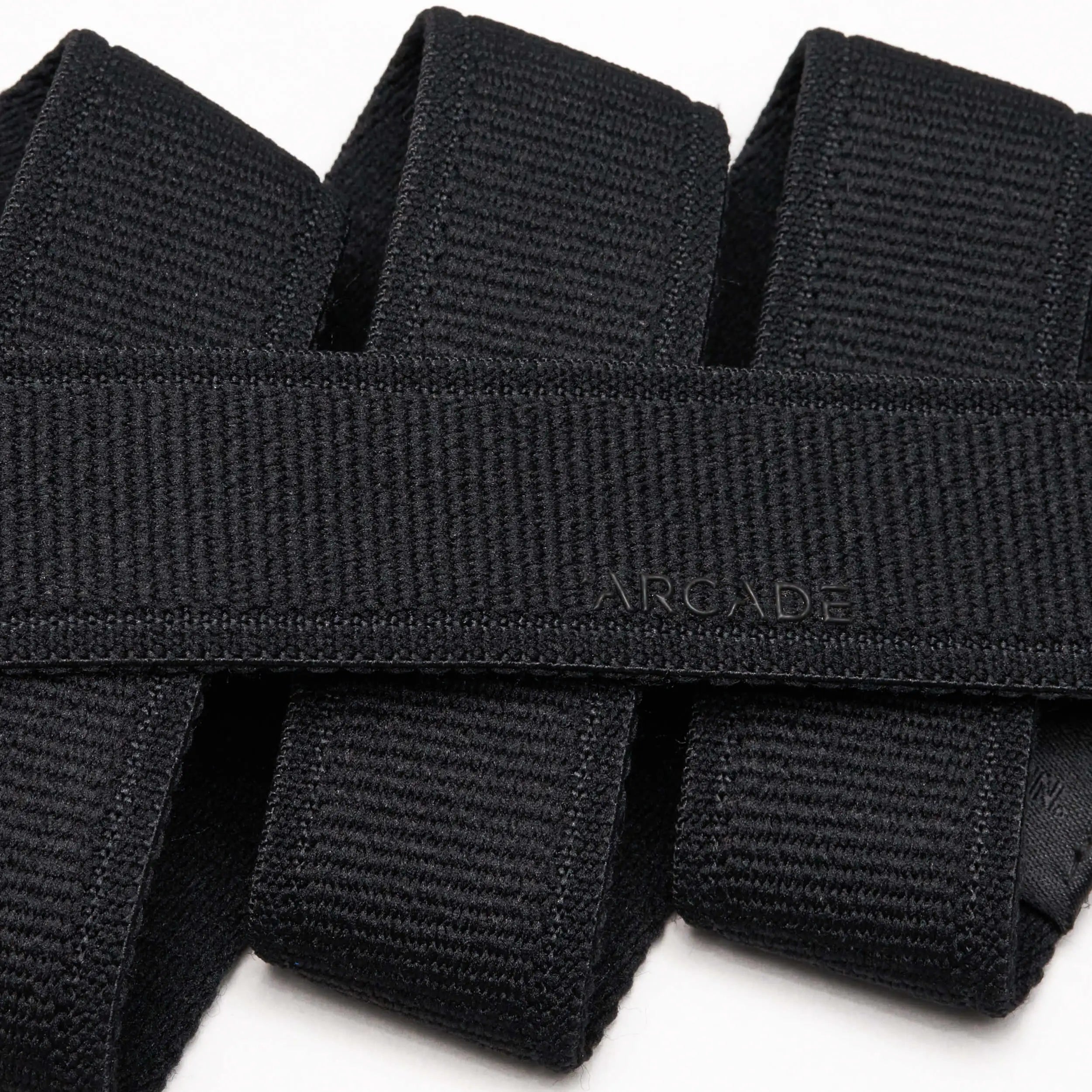A close-up photo of overlapping black Atlas belts from Arcade Belts, with the word ARCADE subtly embossed on one of the elastic stretch belts near the center.