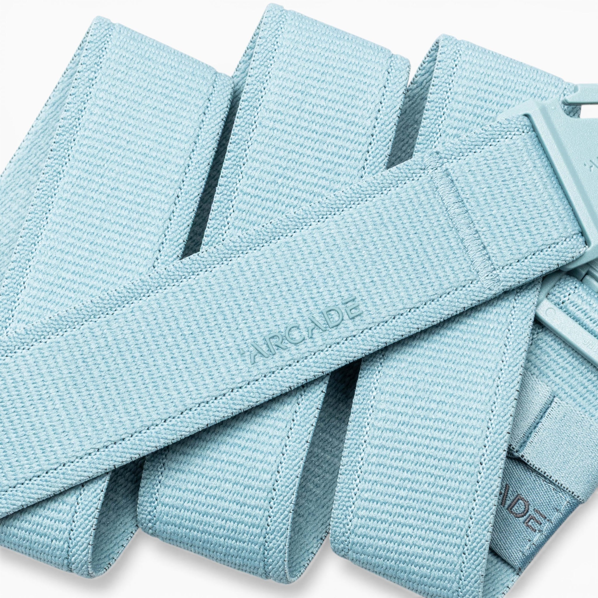 A close-up of the Atlas adventure belt by Arcade Belts in light blue, featuring a plastic A2 buckle. The word “ARCADE” is embossed on the belt’s fabric. This performance stretch belt is coiled and layered over itself.