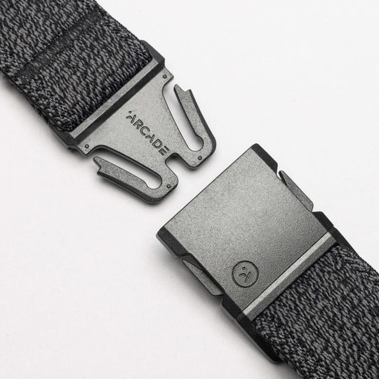 A close-up of the gray woven Atlas belt by Arcade Belts, featuring a black buckle engraved with ARCADE. The partially unclasped buckle uses a simple, secure fastening mechanism—ideal for travel.