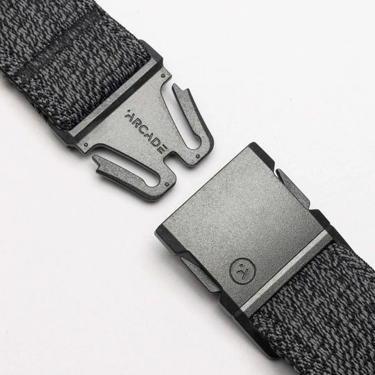 A close-up of the gray woven Atlas belt by Arcade Belts, featuring a black buckle engraved with ARCADE. The partially unclasped buckle uses a simple, secure fastening mechanism—ideal for travel.