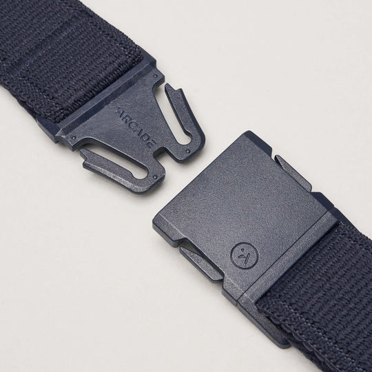 A close-up of a black Atlas belt by Arcade Belts with a plastic buckle, showing the separated ends—one featuring ARCADE, the other a small sad face icon—highlighting its stretch webbing design on a light background.