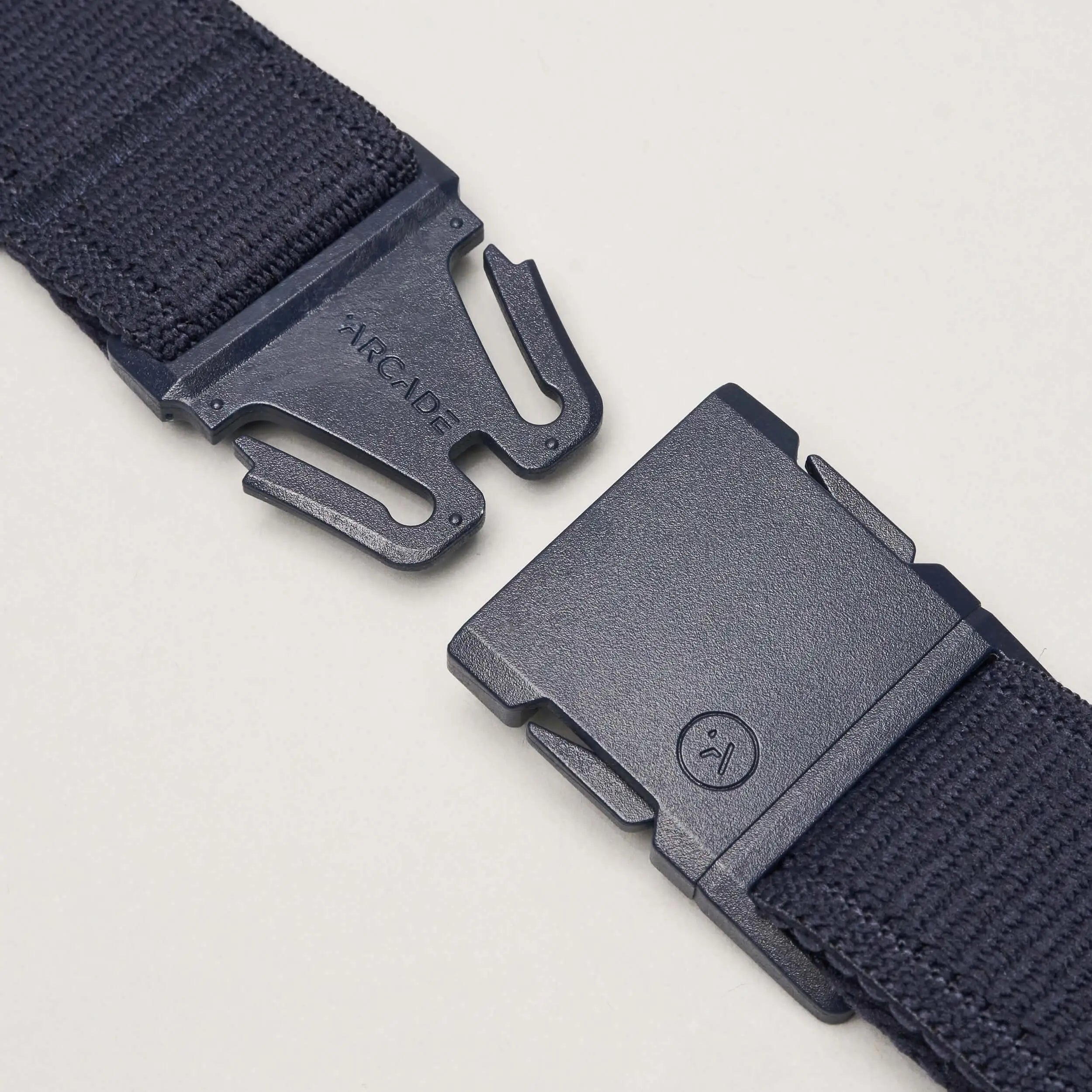 A close-up of a black Atlas belt by Arcade Belts with a plastic buckle, showing the separated ends—one featuring ARCADE, the other a small sad face icon—highlighting its stretch webbing design on a light background.