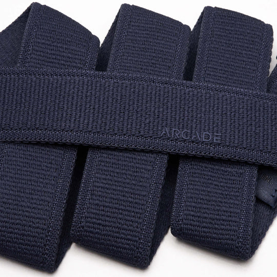 Close-up of several folded navy blue Atlas stretch webbing belts by Arcade Belts stacked neatly, with one low profile belt subtly embossed with ARCADE on its surface.