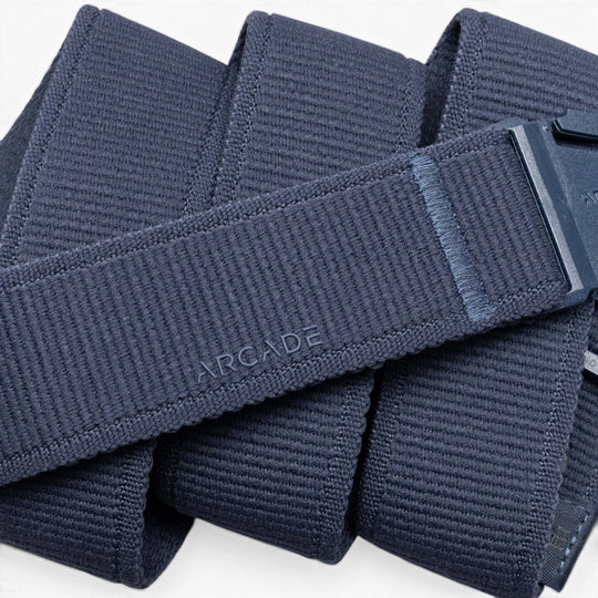 A close-up of the Atlas by Arcade Belts shows its navy blue stretch webbing, plastic buckle with ARCADE embossed near it, and the belt coiled in overlapping loops.
