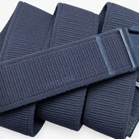 A close-up of the Atlas by Arcade Belts shows its navy blue stretch webbing, plastic buckle with ARCADE embossed near it, and the belt coiled in overlapping loops.