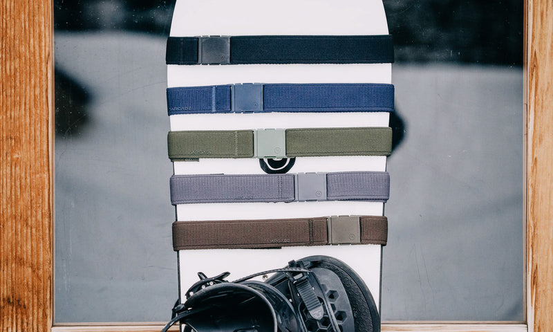 Five different colored stretch webbing travel belts with rectangular buckles are displayed horizontally on a white snowboard, which is standing upright. A black snowboard binding is visible at the bottom of the image.