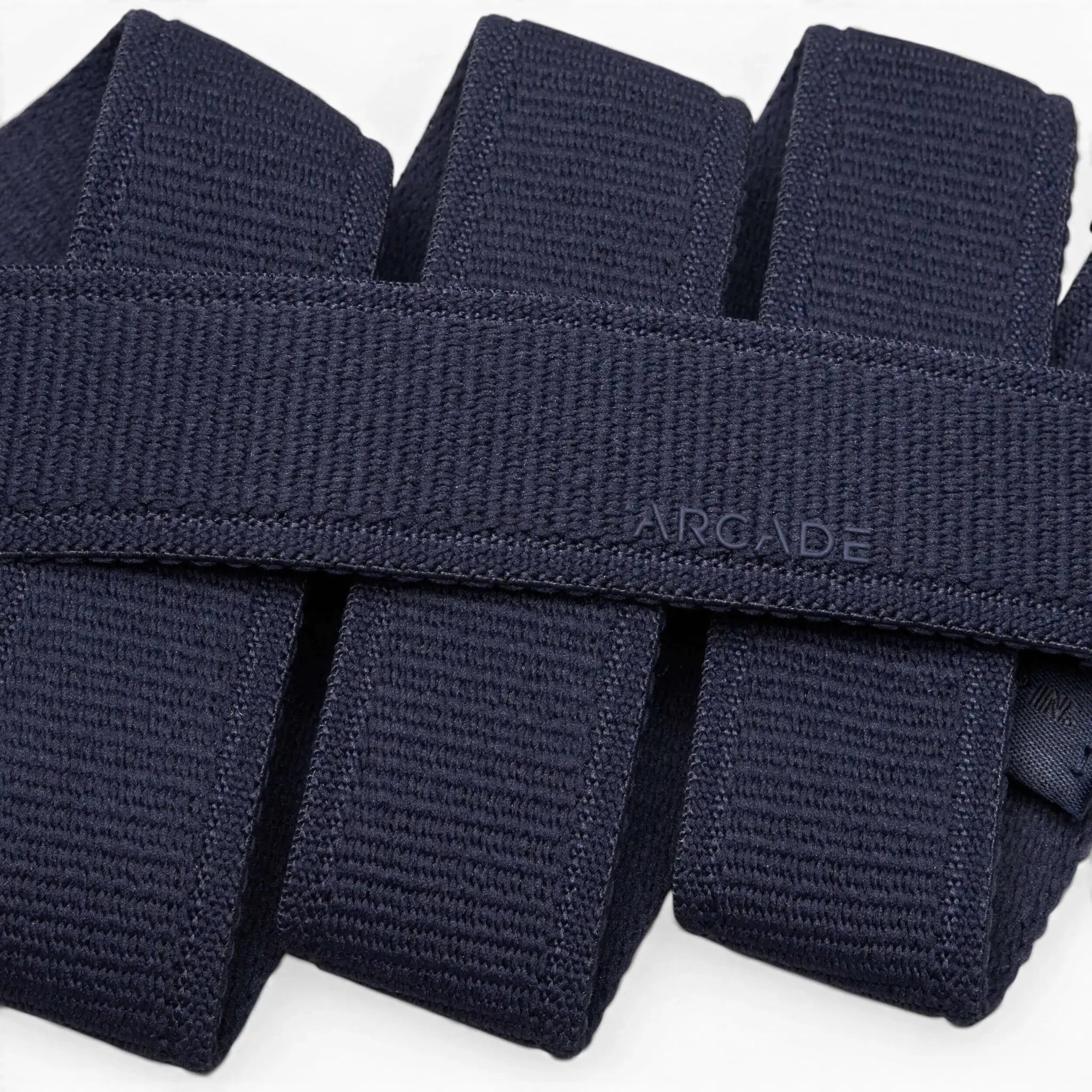 Close-up of several folded navy blue Atlas stretch webbing belts by Arcade Belts stacked neatly, with one low profile belt subtly embossed with ARCADE on its surface.