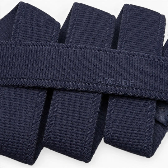 Close-up of several folded navy blue Atlas stretch webbing belts by Arcade Belts stacked neatly, with one low profile belt subtly embossed with ARCADE on its surface.