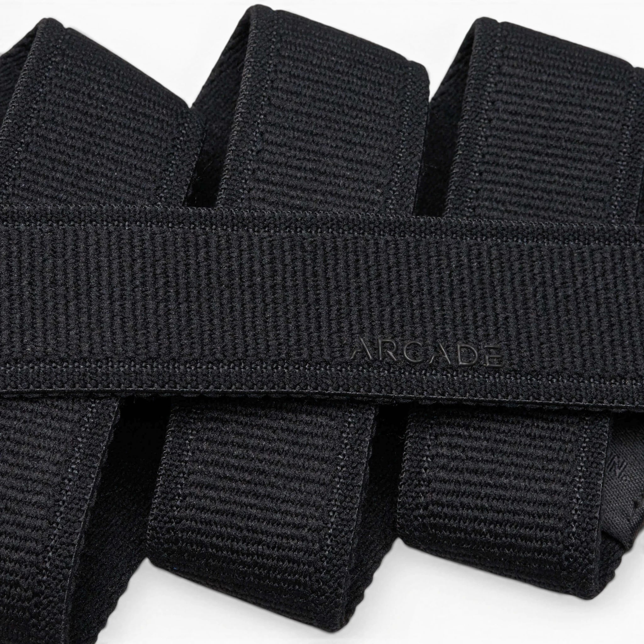A close-up photo of overlapping black Atlas belts from Arcade Belts, with the word ARCADE subtly embossed on one of the elastic stretch belts near the center.