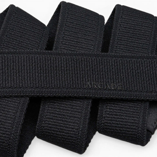 A close-up photo of overlapping black Atlas belts from Arcade Belts, with the word ARCADE subtly embossed on one of the elastic stretch belts near the center.