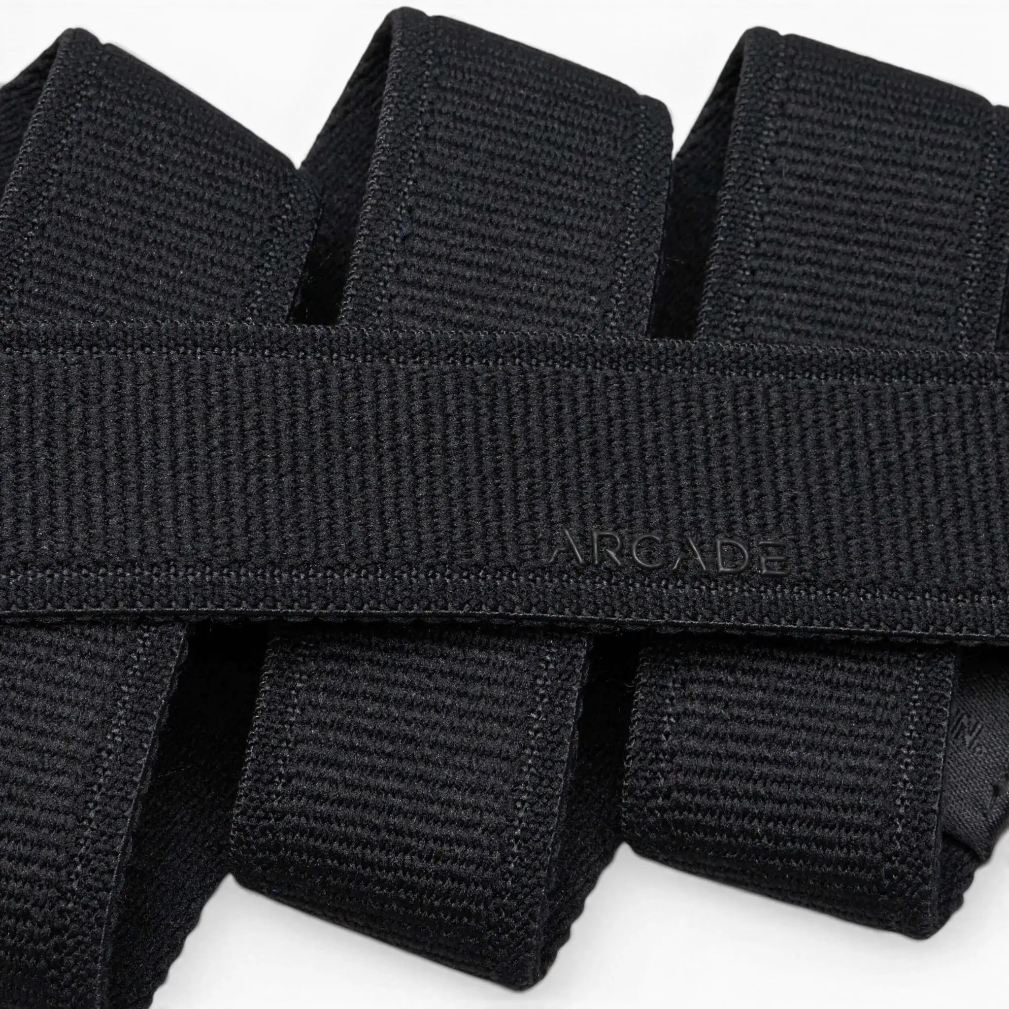 A close-up photo of overlapping black Atlas belts from Arcade Belts, with the word ARCADE subtly embossed on one of the elastic stretch belts near the center.