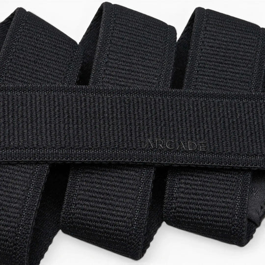 A close-up photo of overlapping black Atlas belts from Arcade Belts, with the word ARCADE subtly embossed on one of the elastic stretch belts near the center.