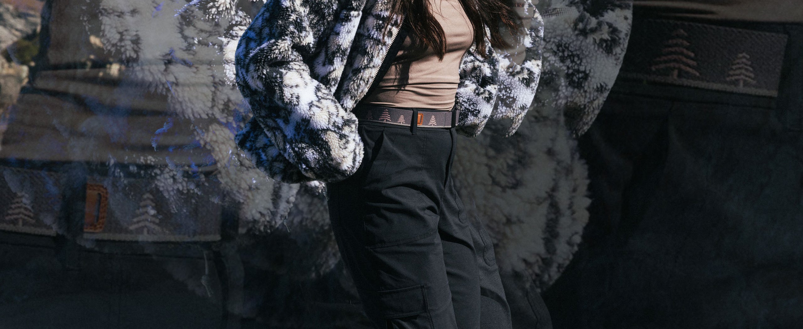 A person wearing a fuzzy black and white jacket, a beige top, and black pants with a belt featuring small tree symbols, standing outdoors with a blurred background.