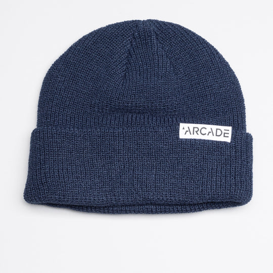 The Arcade Cuff Beanie by Arcade Belts is a blue knit hat with a folded brim and a white rectangular patch displaying ARCADE in black letters on the front, from the creators of innovative elastic stretch belts.