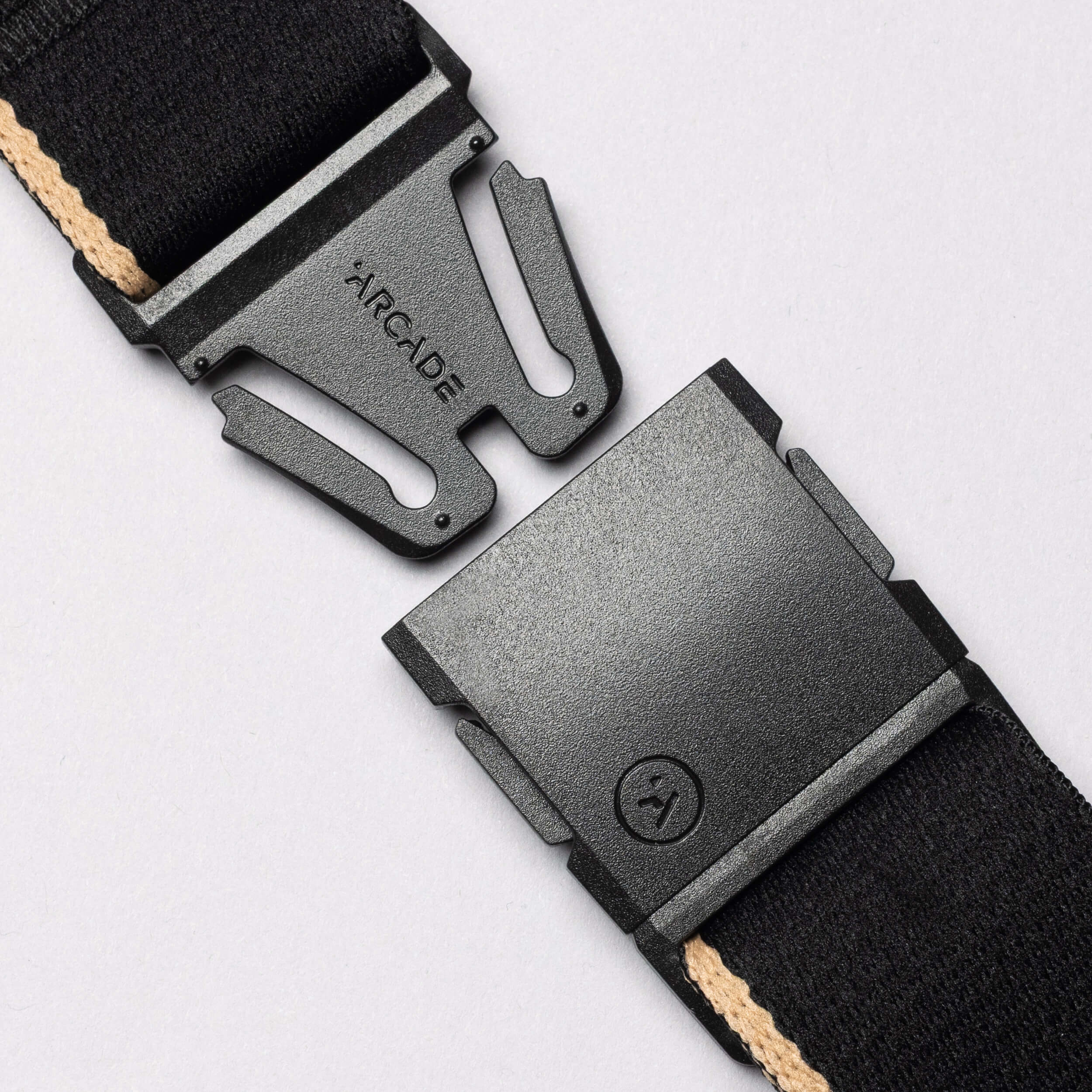 Close-up of the Arcade Belts Blackwood: a black plastic A2 buckle with ARCADE embossed, partially unclasped, attached to a black strap with beige stripe—evoking Blackwood trees—set against a light gray background.