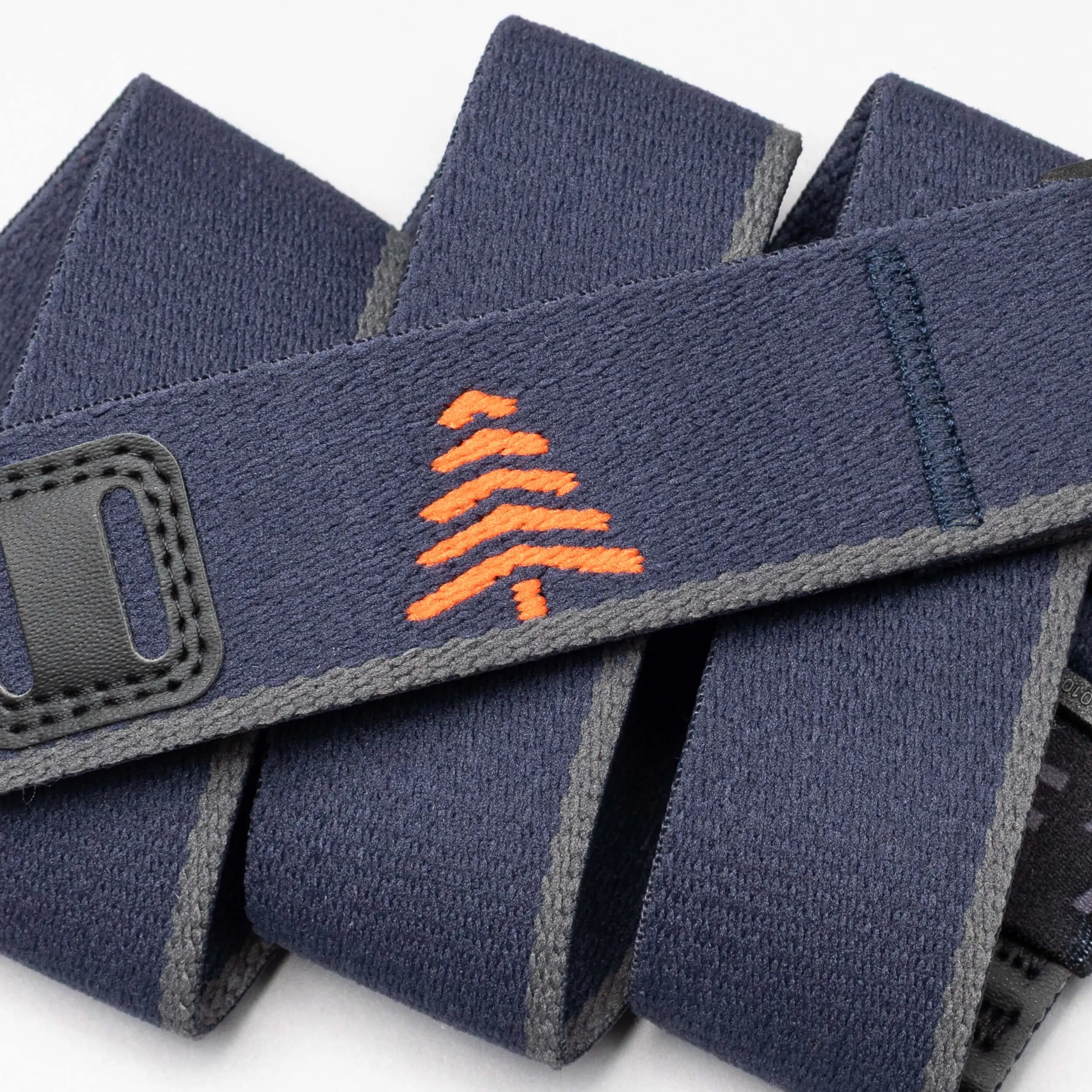 A close-up of the Arcade Belts Blackwood shows a navy blue strap with grey edges, an orange embroidered tree in the center, and black stitched accents, showcasing its stylish craftsmanship.