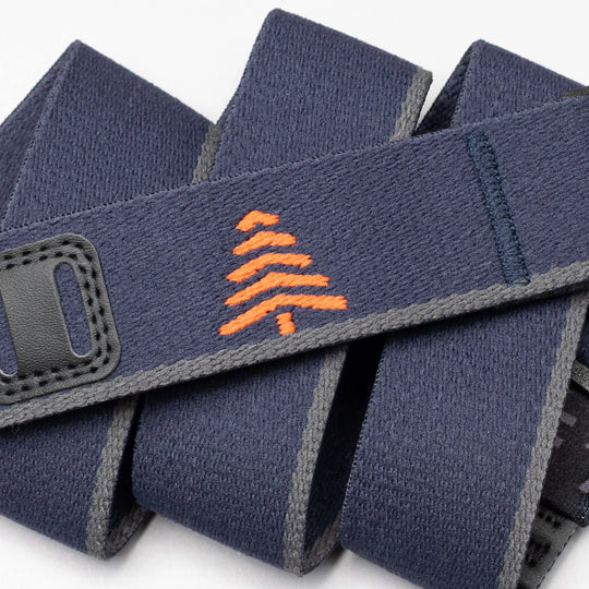 A close-up of the Arcade Belts Blackwood shows a navy blue strap with grey edges, an orange embroidered tree in the center, and black stitched accents, showcasing its stylish craftsmanship.