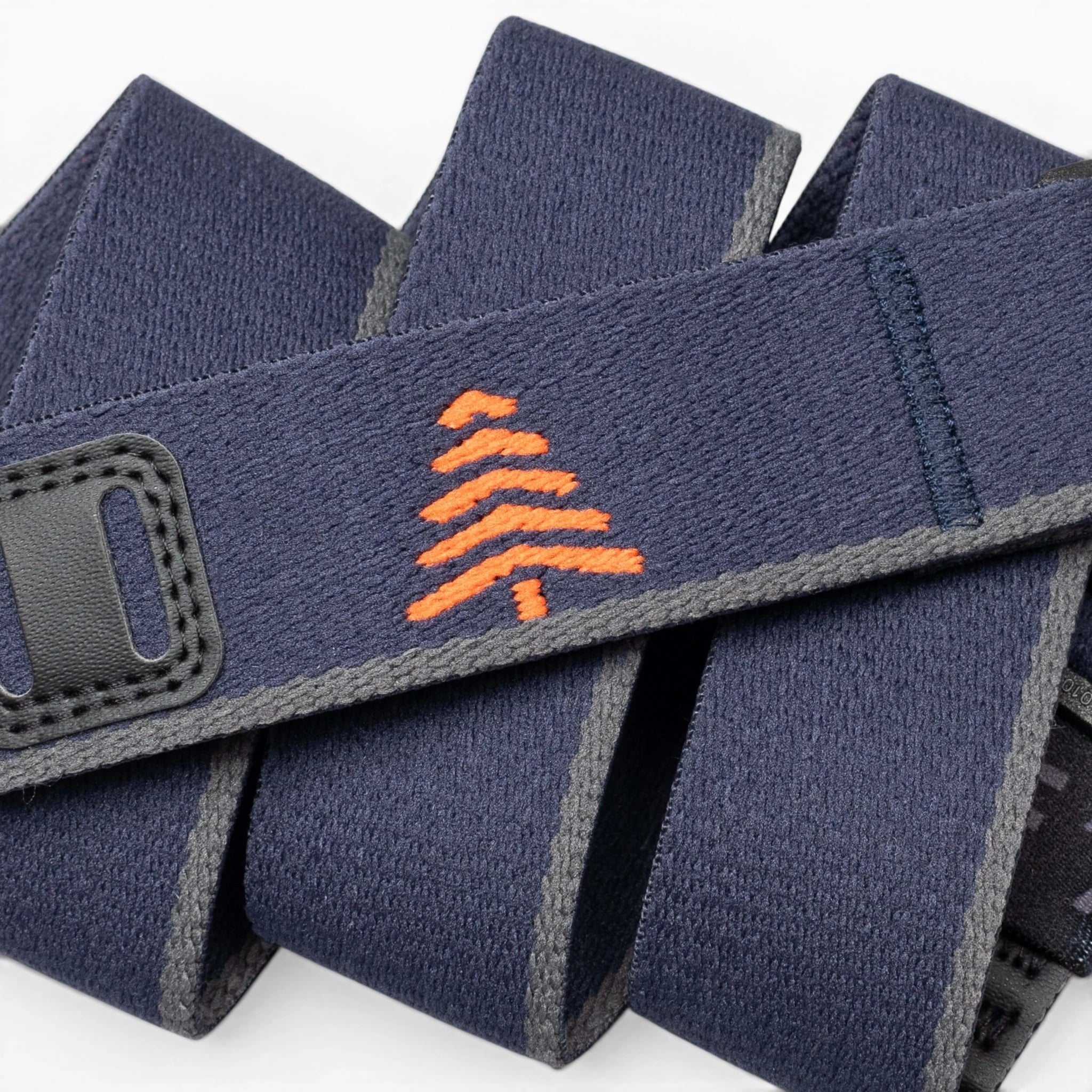 A close-up of the Arcade Belts Blackwood shows a navy blue strap with grey edges, an orange embroidered tree in the center, and black stitched accents, showcasing its stylish craftsmanship.