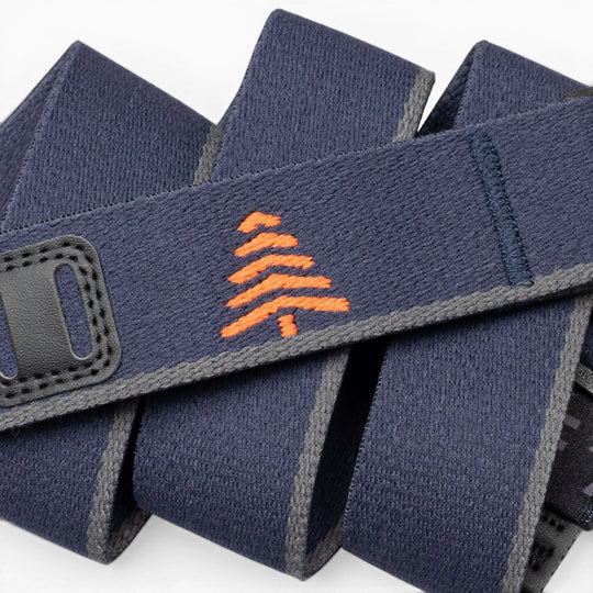 A close-up of the Arcade Belts Blackwood shows a navy blue strap with grey edges, an orange embroidered tree in the center, and black stitched accents, showcasing its stylish craftsmanship.